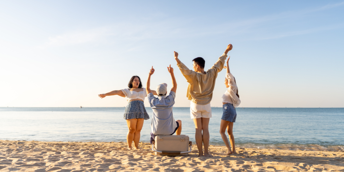 Group of friends celebrating with arms raised facing the ocean on a peaceful beach at sunset.