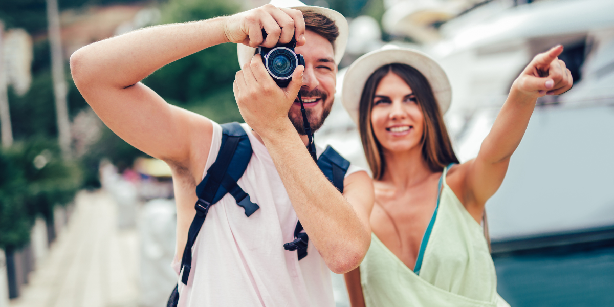 Pareja de turistas sonrientes en una marina; hombre tomando foto con cámara DSLR y mujer señalando al frente.