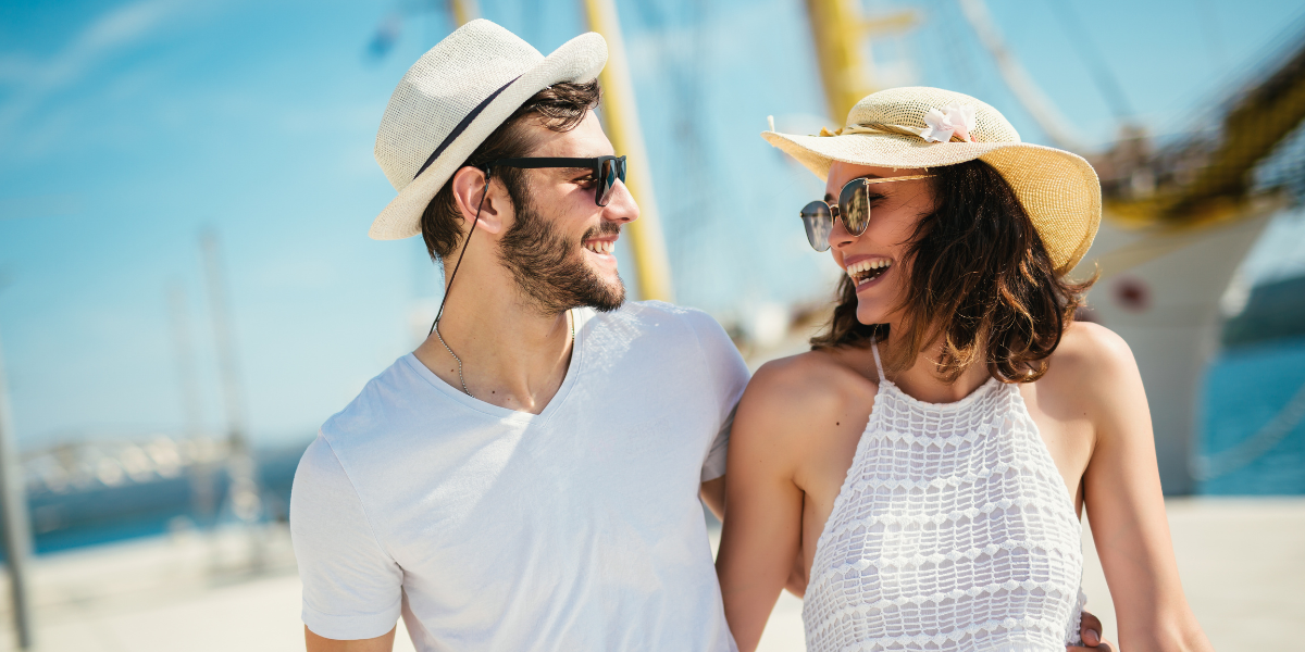 Young couple wearing hats and sunglasses smiling at a marina with yachts in the background.