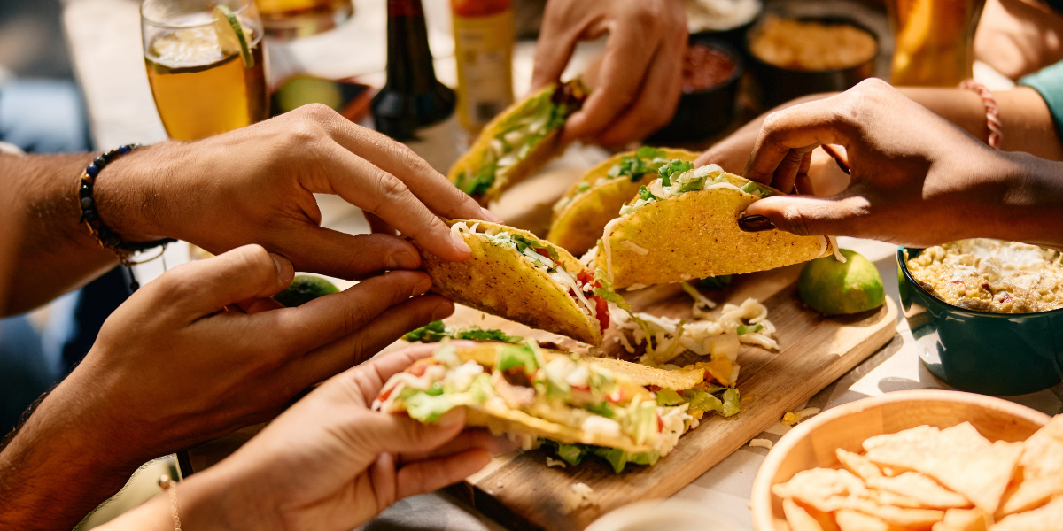 Close-up of hands sharing Mexican corn tortilla tacos with fresh vegetables and salsas.