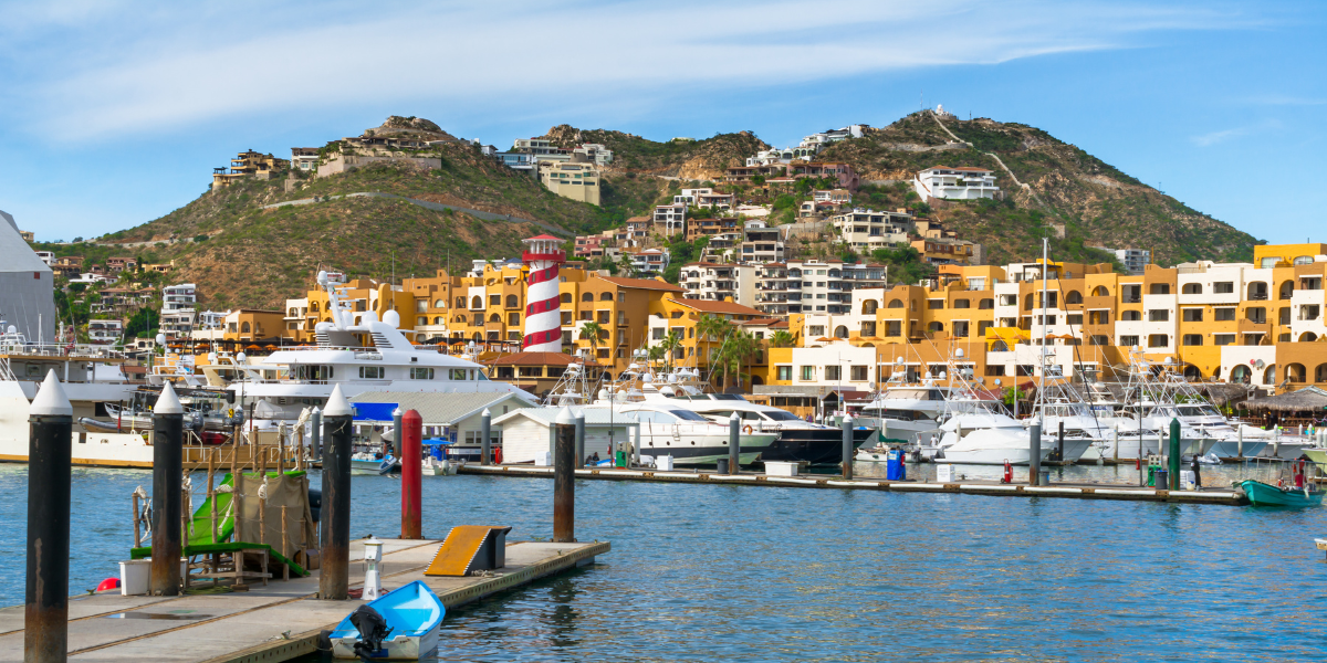 Puerto deportivo de Cabo San Lucas con yates de lujo, edificios coloridos y faro a rayas rojas y blancas.