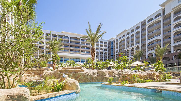 Resort pool with turquoise water, stone features, palm trees, and multi-story hotel building.